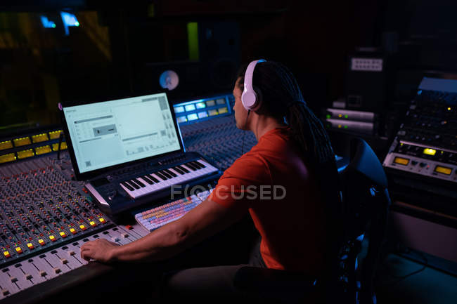 Seitenansicht eines jungen männlichen Tontechnikers, der an einem Mischpult in einem Tonstudio sitzt und mit einem Computer arbeitet und Kopfhörer trägt — Stockfoto