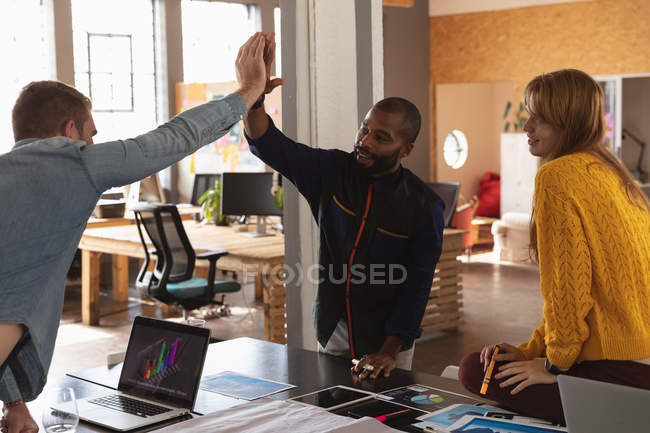 Front view close up of a young African American man and a young Caucasian man high fiving over a desk and a female Caucasian colleague sitting on a desk smiling in a creative office — Stock Photo
