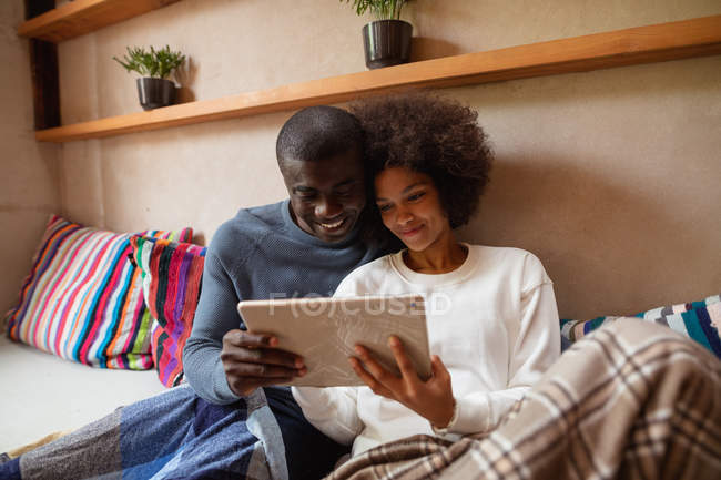 Vista frontal de cerca de una joven mestiza y un joven afroamericano mirando una tableta y sonriendo, sentados juntos en un sofá en casa . - foto de stock