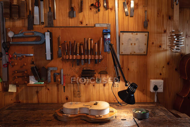 Violín hecho en un taller más luthier con herramientas colgando en la pared en el fondo - foto de stock
