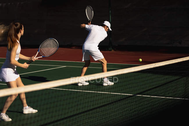 Вид сбоку молодая кавказка и мужчина, играющий в теннис в солнечный день, мужчина, возвращающий мяч — стоковое фото