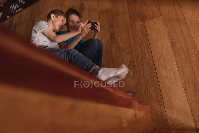 Погляд збоку на двох дошкільнят, які сидять вдома на сходах, за допомогою смартфона. — стокове фото