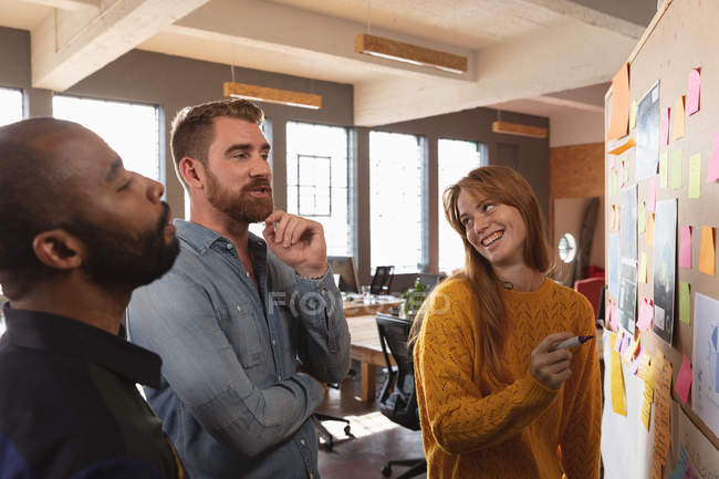 Side view close up of a young African American man and a young Caucasian man and woman standing talking and writing on an ideas board during a team brainstorm session at a creative office — Stock Photo