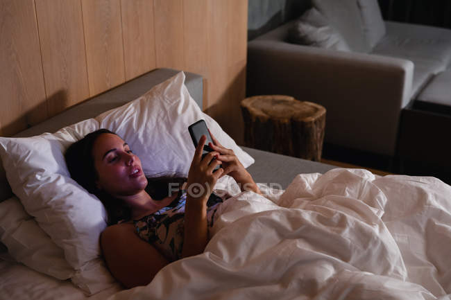 Erhöhte Ansicht einer jungen kaukasischen brünetten Frau, die mit einem Smartphone auf dem Rücken im Bett liegt — Stockfoto