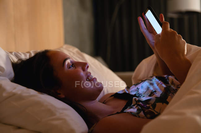 Side view close up of a smiling young Caucasian brunette woman lying on her back in bed using a smartphone — Stock Photo