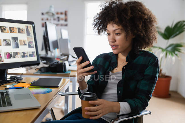 Vista lateral de perto de uma jovem mulher de raça mista sentada em uma mesa e usando um smartphone em um escritório criativo — Fotografia de Stock