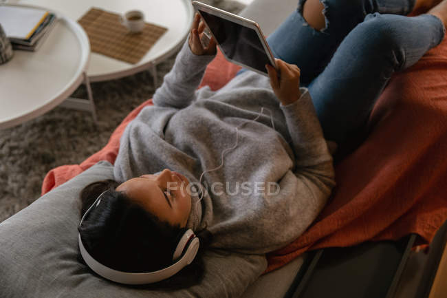 Підвищений вид молодий Кавказький жінка брюнетка лежачи на спині на дивані, носіння навушників і перегляду планшетного комп'ютера — стокове фото