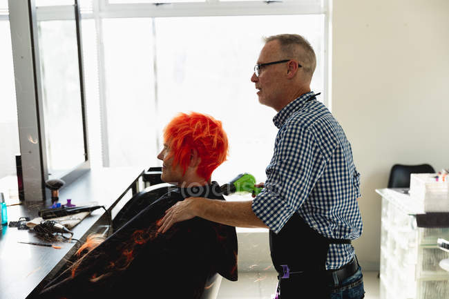 Vista lateral de um cabeleireiro masculino caucasiano de meia idade e uma jovem mulher caucasiana com seu cabelo colorido vermelho brilhante e golpe seco em um salão de cabeleireiro — Fotografia de Stock
