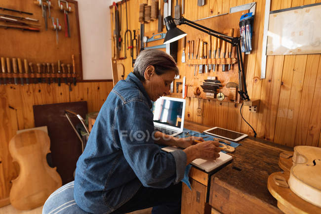 Side view of a senior Caucasian female luthier making notes in her workshop, with a laptop and a tablet computer in front of her and tools hanging up on the wall in the background — Stock Photo