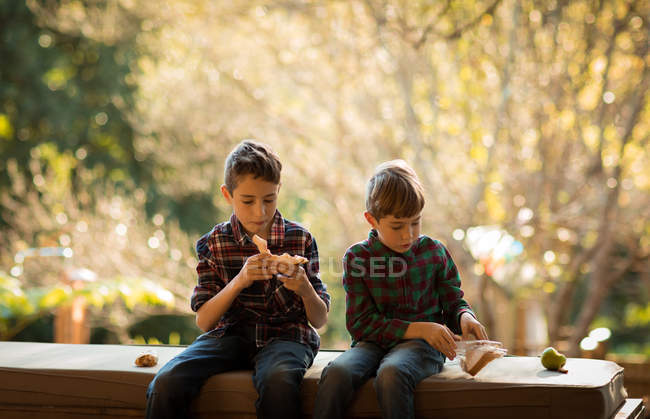 Vista frontal de dos niños caucásicos pre adolescentes sentados en un banco en un jardín, almorzando - foto de stock