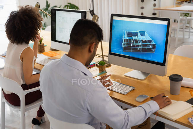 Вид ззаду крупним планом молодий чоловік змішаної раси і молода змішана жінка, що сидить за столом, використовуючи комп'ютери в творчому офісі — стокове фото