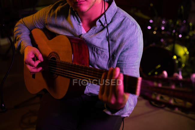 Vista frontal sección media de un joven caucásico sentado y tocando una guitarra acústica durante una sesión en un estudio de grabación - foto de stock