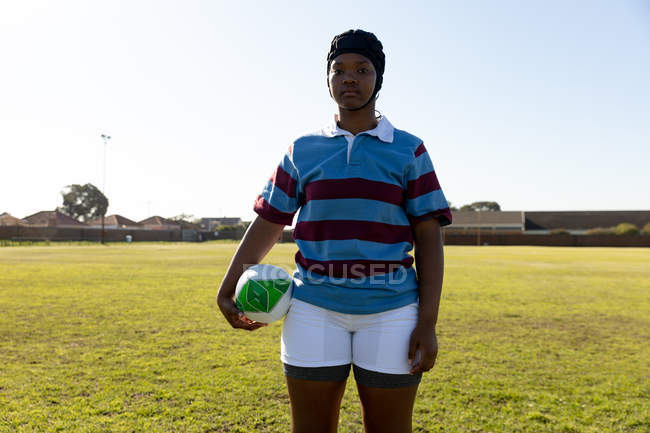 Портрет молодой взрослой регбистки смешанной расы, одетой в охранника, стоящего на регби, держащего мяч для регби, смотрящий в камеру — стоковое фото