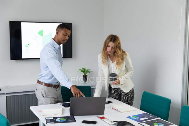 Vista frontal de un joven afroamericano y una joven mujer caucásica de pie y hablando en un escritorio usando una computadora portátil en la oficina moderna de un negocio creativo, el hombre señala a la pantalla y la mujer sostiene un café para llevar - foto de stock