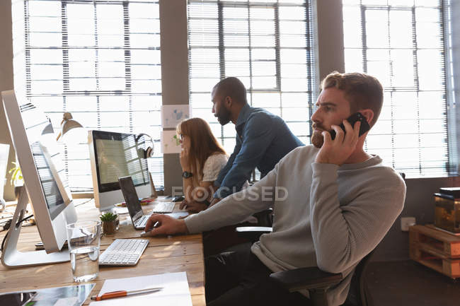 Вид сбоку на молодого кавказца, сидящего за столом, разговаривающего на смартфоне в креативном офисе, с двумя коллегами, смотрящими на монитор и на заднем плане — стоковое фото