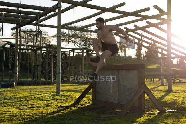 Vista lateral de um jovem caucasiano sem camisa saltando sobre uma parede em um ginásio ao ar livre durante uma sessão de treinamento de bootcamp — Fotografia de Stock