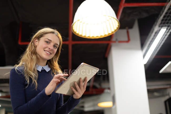 Vista frontale da vicino di una giovane donna caucasica in piedi utilizzando un tablet, guardando la fotocamera sorridente nel moderno ufficio di un business creativo — Foto stock