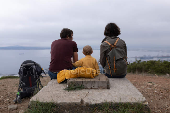 Задний вид на молодого кавказского отца и мать, сидящих в парке с ребенком между ними и наслаждающихся видом — стоковое фото