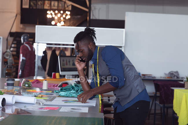 Side view of a young African American male fashion student talking on a smartphone and working on a design in a studio at fashion college — Stock Photo