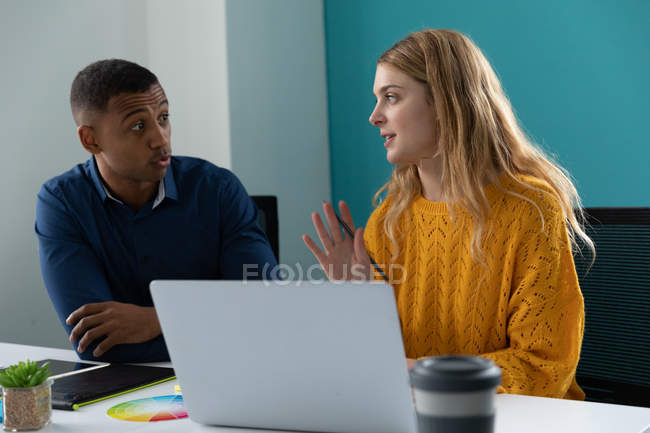 Vista frontal de perto de um jovem afro-americano ouvindo e uma jovem caucasiana usando um computador portátil e conversando, sentados juntos em uma mesa no escritório moderno de um negócio criativo — Fotografia de Stock