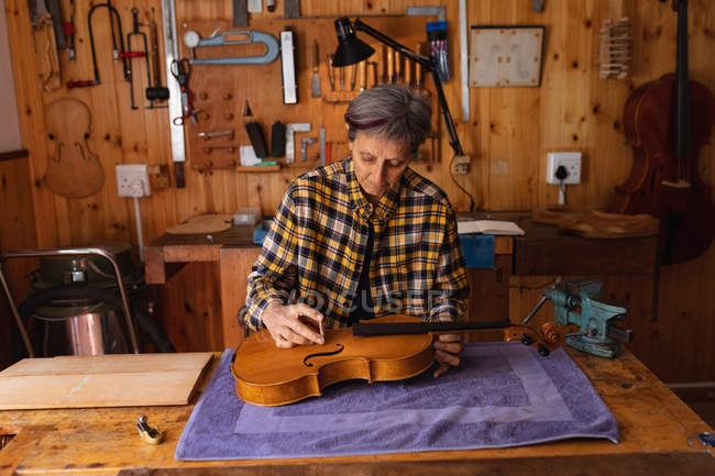 Vorderansicht einer älteren kaukasischen Geigenbauerin, die in ihrer Werkstatt am Körper einer Geige arbeitet, im Hintergrund hängen Werkzeuge an der Wand — Stockfoto