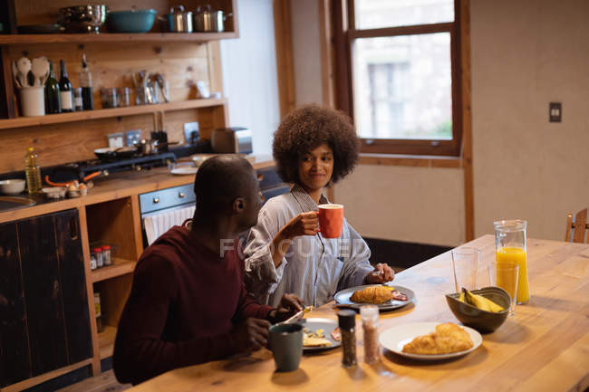 Vista frontal de uma jovem mulher de raça mista sorridente e seu parceiro, um jovem afro-americano, comendo, conversando e bebendo café sentado em sua mesa de cozinha em casa — Fotografia de Stock