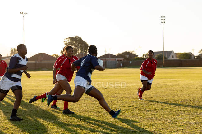 Vista lateral de un grupo de jóvenes adultas multi-étnicas jugadoras de rugby corriendo en el campo durante un partido y persiguiendo al jugador en posesión de la pelota - foto de stock