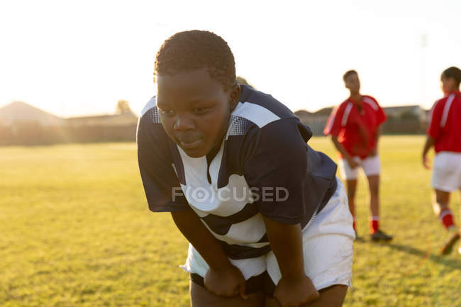 Вид спереди на молодую взрослую афроамериканку, играющую в регби, отдыхающую на поле для регби после матча, с игроками из другой команды на заднем плане — стоковое фото