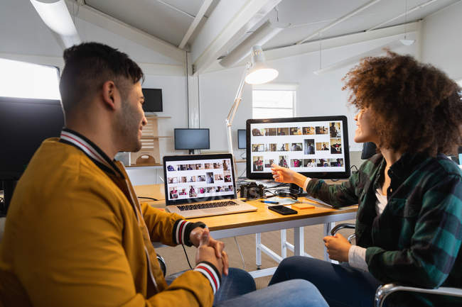 Вид сбоку на молодого человека смешанной расы и молодую женщину смешанной расы, сидящую за столом и смотрящую на экран компьютера и обсуждающую в творческом офисе — стоковое фото