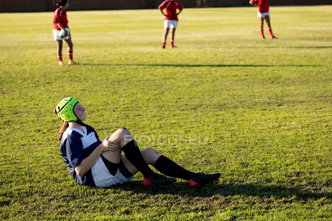 Vista lateral de una joven jugadora de rugby caucásica con un protector de cabeza sentado en un campo de rugby sosteniendo su pierna lesionada, con otros jugadores en el fondo - foto de stock