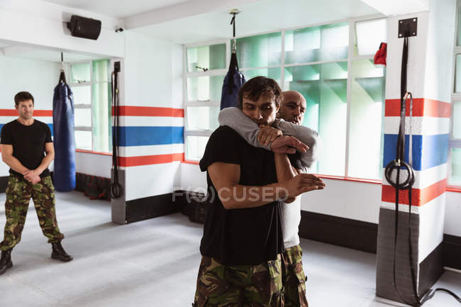 Vue de face d'un instructeur caucasien d'âge moyen qui donne un entraînement d'autodéfense dans un gymnase de boxe démontrant son emprise sur un jeune homme de race mixte, tandis qu'un autre jeune homme regarde — Photo de stock