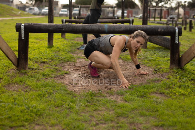 Vista frontal de una joven mujer caucásica arrastrándose bajo un bajo obstáculo en un gimnasio al aire libre durante una sesión de entrenamiento de bootcamp - foto de stock
