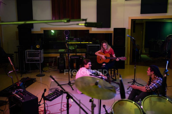 Vista elevada de una joven mujer caucásica sentada en un taburete con una guitarra acústica mientras un joven hombre caucásico y un joven mestizo se sientan en el suelo hablando, durante una sesión de composición de la banda en un estudio de grabación - foto de stock