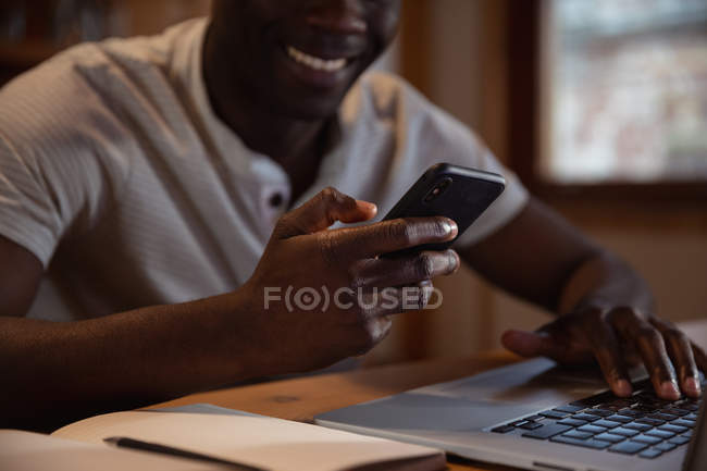 Vista frontal sección central de un joven afroamericano sonriente usando un teléfono inteligente y una computadora portátil sentada en una mesa en casa - foto de stock