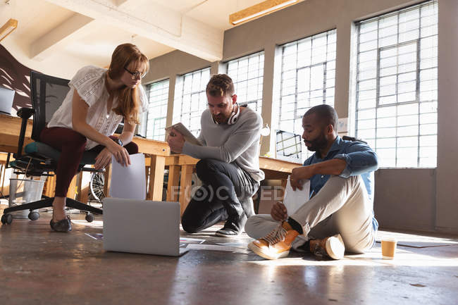Front view close up of a young African American man and a young Caucasian man sitting on the floor and a young Caucasian female colleague sitting on a chair looking at paperwork together — Stock Photo