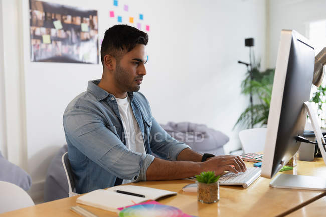 Seitenansicht eines jungen Mannes mit gemischter Rasse, der an einem Schreibtisch sitzt und einen Computer in einem kreativen Büro benutzt — Stockfoto