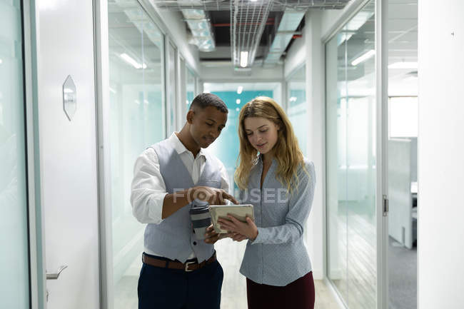 Вид спереди на молодую кавказку и молодого афроамериканца, стоящего и пользующегося планшетным компьютером в коридоре современного офиса — стоковое фото