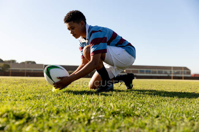 Vue de face rapprochée d'une jeune joueuse de rugby mixte adulte agenouillée sur un terrain de rugby et mettant la balle sur un tee-shirt pour un coup de pied — Photo de stock