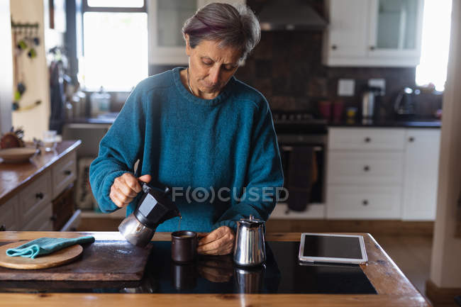 Front view of a senior Caucasian woman in a kitchen pouring coffee with a tablet next to her and cupboards in the background — Stock Photo