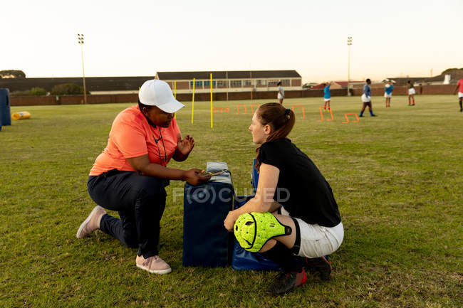 Vista lateral de uma mista de meia-idade treinador de rugby feminino ajoelhado e conversando com uma jovem adulta branca jogadora de rugby feminina em um campo de rugby durante uma sessão de treinamento — Fotografia de Stock
