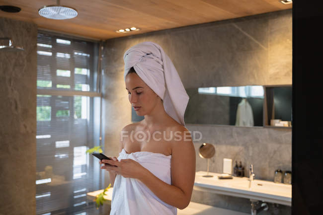 Vista lateral de una joven morena caucásica vestida con una toalla de baño y con el pelo envuelto en una toalla, usando un smartphone en un baño moderno - foto de stock