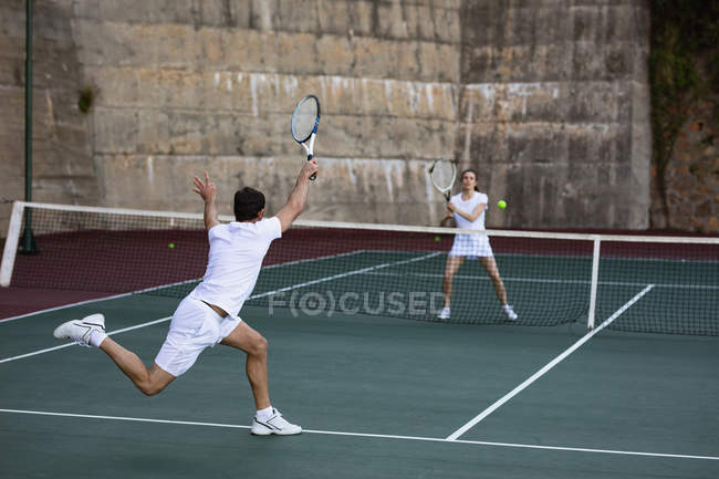 Visão traseira de uma jovem caucasiana e um homem jogando tênis, homem retornando a bola com uma parede no fundo — Fotografia de Stock