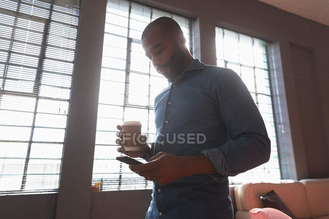 Vue de côté gros plan d'un jeune Afro-Américain tenant un café à emporter et utilisant un smartphone debout devant une fenêtre dans un bureau créatif, rétroéclairé par la lumière du soleil — Photo de stock