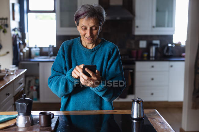 Vista frontal de uma mulher caucasiana sênior em uma cozinha usando um smartphone com armários de cozinha ao fundo — Fotografia de Stock