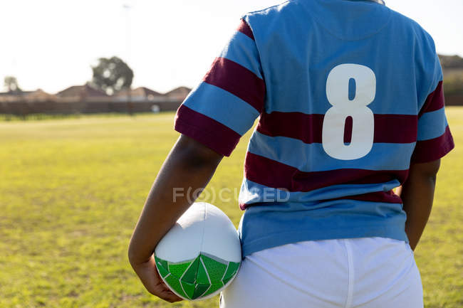Visão traseira seção média do jogador de rugby feminino em pé em um campo de rugby com uma bola de rugby sob seu braço — Fotografia de Stock