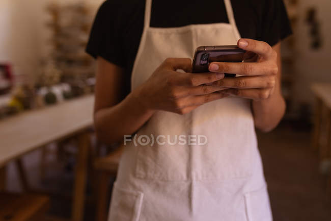 Vue de face du milieu d'un potier féminin portant un tablier à l'aide d'un téléphone intelligent dans un atelier de poterie — Photo de stock