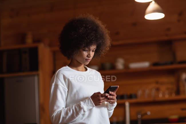 Vista frontal close-up de uma jovem mulher de raça mista em pé em sua cozinha usando um smartphone — Fotografia de Stock