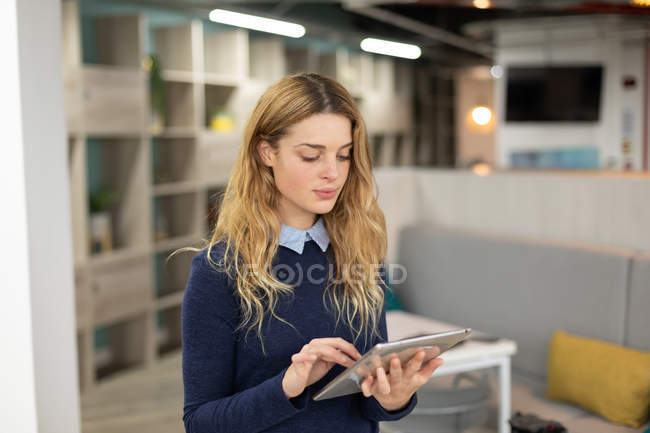 Vista frontale di una giovane donna caucasica in piedi utilizzando un computer tablet nella zona lounge di un moderno business creativo — Foto stock