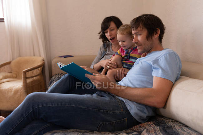 Vista laterale di un giovane padre e madre caucasici seduti su un divano e che leggono un libro con il loro bambino — Foto stock