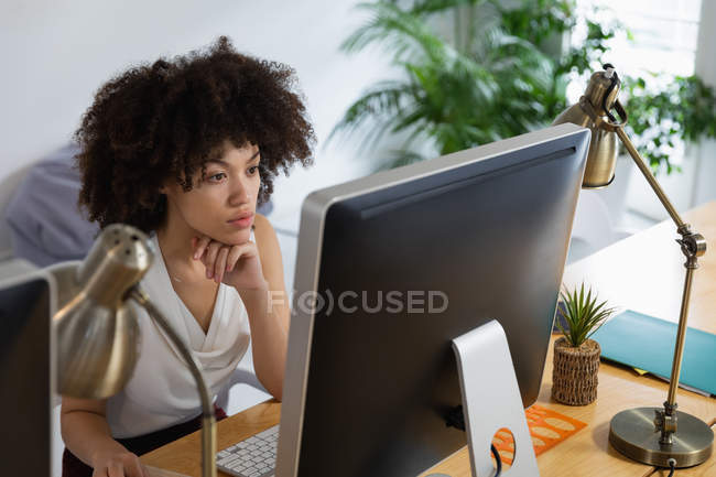 Vista frontal de uma jovem mulher de raça mista sentada em uma mesa olhando para uma tela de computador em um escritório criativo — Fotografia de Stock